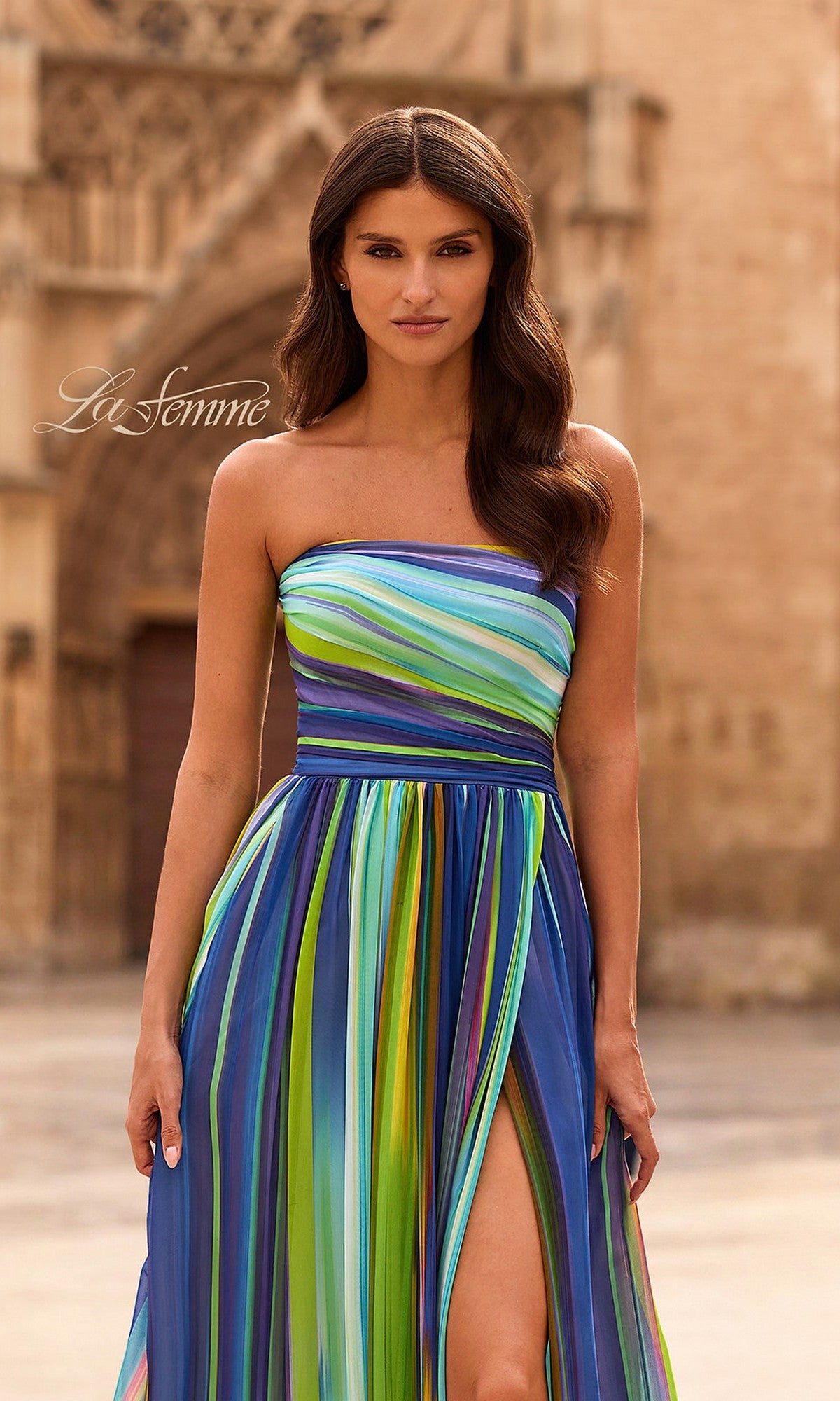 A woman wearing the La Femme 33690 Multi-Colored Prom Dress, featuring multicolored stripes and a thigh-high slit, stands outdoors in front of a stone building.